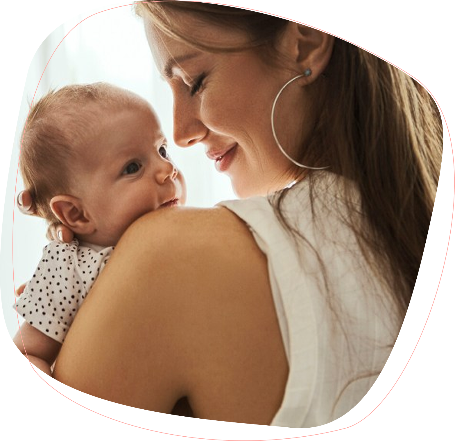 A woman with long brown hair smiles gently while holding a baby in a white outfit with black dots, resting the baby on her shoulder. The baby looks up at her, both appearing content and close.