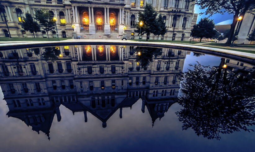 Elegant historic building with glowing lights reflected clearly in a large, still pool of water at dusk, with surrounding trees and part of the sky also visible in the reflection.