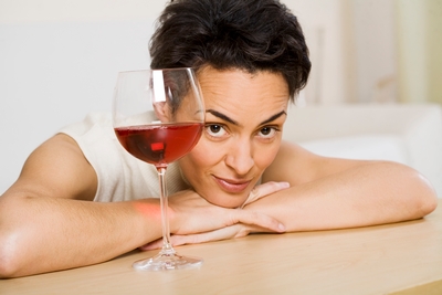 A person with short dark hair rests their chin on their hands on a table, looking at the camera. A glass of red wine sits on the table in front of them.