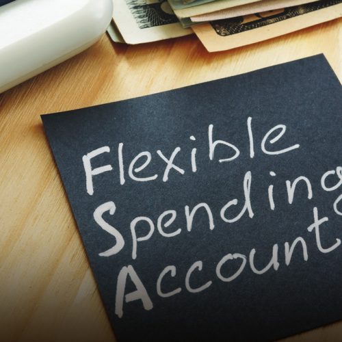 A black sticky note with Flexible Spending Account written on it sits on a wooden desk near a calculator and a stack of cash.