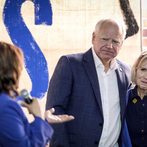 A woman holding a microphone speaks to a man and woman standing together. The man has his arm around the woman, and both are wearing blue jackets. A large sign with the letters IS is visible in the background.