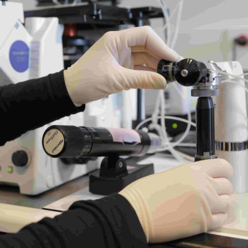 A person wearing gloves adjusts scientific equipment in a laboratory setting, surrounded by various instruments and cables on a workbench.
