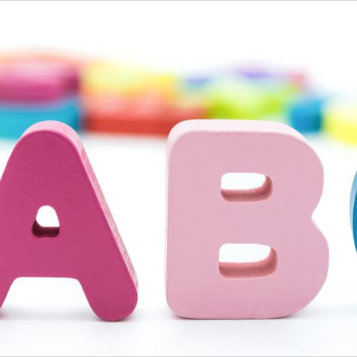 Three colorful wooden letters—A in red, B in pink, and C in blue—stand upright in the foreground, with blurred multicolored shapes in the background on a white surface.