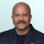A smiling man with a mustache and beard, wearing a dark blue blazer, matching sweater, and a blue pocket square, poses in front of a light blue background.