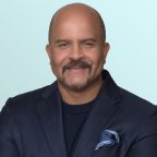 A smiling man with a mustache and beard, wearing a dark blue blazer, matching sweater, and a blue pocket square, poses in front of a light blue background.