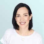 A woman with straight dark hair and light skin smiles at the camera. She is wearing a white shirt and a delicate gold necklace, and the background is a soft, light blue gradient.