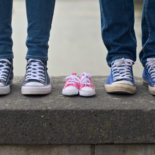Two adults in jeans and sneakers stand on either side of a small pair of pink baby shoes, placed between them on a stone ledge, suggesting an upcoming addition to the family.
