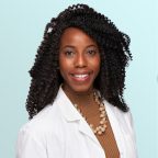 A smiling woman with curly hair wearing a white lab coat, a brown turtleneck, and a pearl necklace, posing in front of a light blue background.