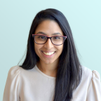 A woman with long dark hair and glasses smiles at the camera. She is wearing a light-colored blouse and is posed in front of a plain light blue background.