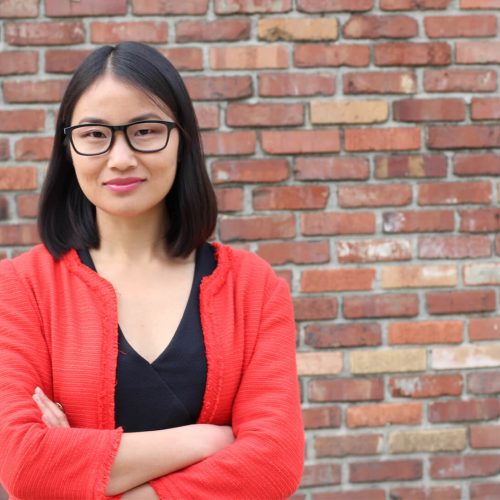 A woman with straight black hair and glasses stands in front of a brick wall, wearing a red jacket over a black top, with her arms crossed and a slight smile.