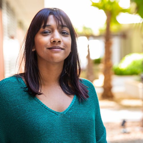 A woman with straight dark hair and bangs, wearing a teal sweater, stands outdoors and smiles slightly. Palm trees and sunlight are visible in the background, creating a warm, relaxed atmosphere.