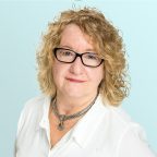 A woman with curly blonde hair and black glasses wearing a white blouse and a chunky silver necklace, posed against a light blue background.