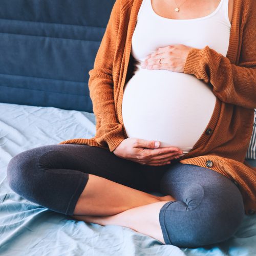 Beautiful pregnant woman sitting at bed and holds hands on belly in bedroom at home. Young mother waiting of a baby. Pregnancy, maternity and expectation image. Close-up, indoors.