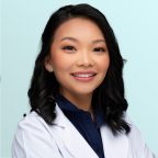 A woman with wavy black hair wearing a white lab coat and navy blouse smiles at the camera against a light blue background.