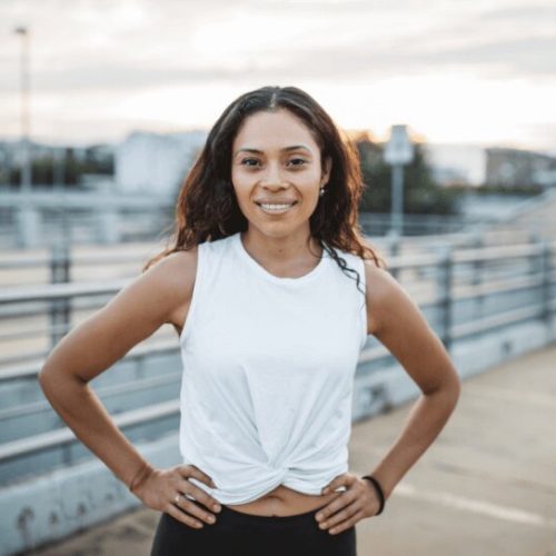 A woman with long curly hair, wearing a white sleeveless top and black leggings, stands outside with her hands on her hips, smiling at the camera. The background shows an urban setting with a railing and buildings.