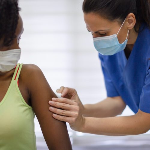 A healthcare worker in blue scrubs and a face mask administers a vaccine to a woman wearing a light green tank top and a mask. The woman looks at her arm as the shot is given.