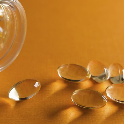 A glass bottle tipped on its side spills several oval, translucent, yellow softgel capsules onto a bright orange surface.