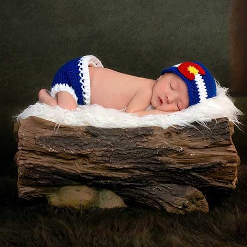 A newborn baby sleeps on a white fuzzy blanket atop a log prop, wearing a blue knit hat with a red and yellow accent and matching blue diaper cover, set against a dark forest-themed background.