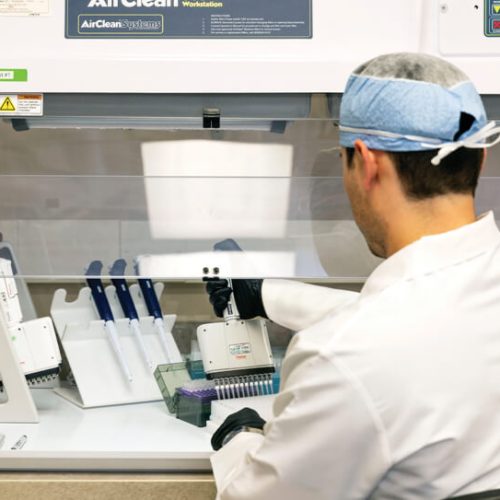 A laboratory technician wearing a lab coat, gloves, and a hair cover works under a fume hood, using a pipette to dispense liquid into a tray with several pipettes and lab equipment nearby.