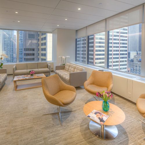 Modern office lounge with tan armchairs, sofas, coffee tables with magazines and flowers, large windows overlooking city skyscrapers, and neutral-toned décor.