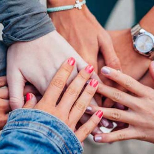 A group of people place their hands together in the center, forming a circle. Their hands overlap, symbolizing unity and teamwork. Some wear watches or bracelets, and a variety of skin tones and nail colors are visible.