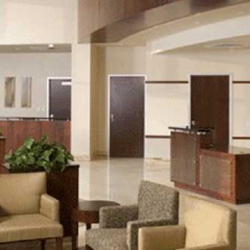 Modern office lobby with beige floors, brown and tan armchairs, potted plants, wood reception desks, and abstract art on the wall, creating a clean and welcoming atmosphere.