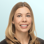 A woman with long, light brown hair and hazel eyes smiles at the camera. She is wearing a grey top and small earrings, with a light blue background behind her.