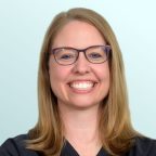 A woman with shoulder-length blonde hair and glasses smiles broadly. She is wearing black medical scrubs with embroidered text on the chest and stands against a light blue background.