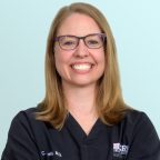 A woman with shoulder-length blonde hair and glasses smiles broadly. She is wearing black medical scrubs with embroidered text on the chest and stands against a light blue background.