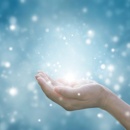 A hand gently cupped and extended against a blue background, surrounded by glowing, ethereal light and sparkling particles, creating a magical or hopeful atmosphere.