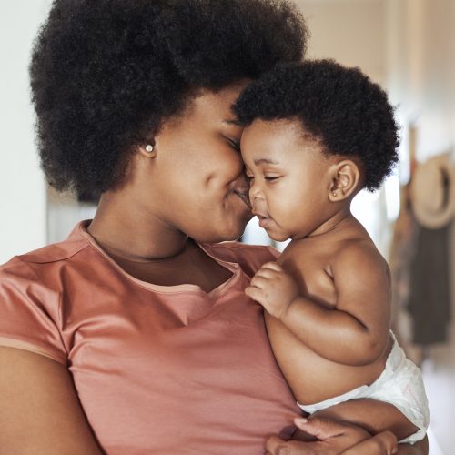 A woman lovingly holds and nuzzles her smiling baby, who is only wearing a diaper. They are indoors, and both share an affectionate moment, their faces close together.