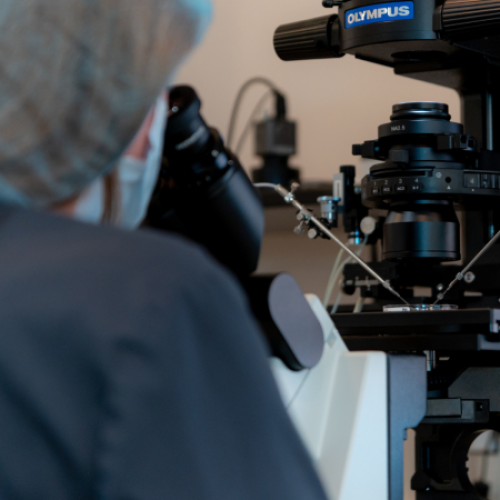 A person wearing a hair cover and lab coat looks through a microscope in a laboratory setting, focusing on scientific equipment with wires and mechanical components.