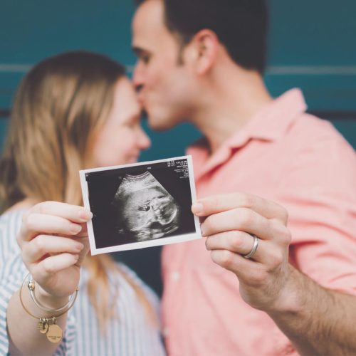 A couple stands close together as the man kisses the woman’s forehead. The woman holds a sonogram photo in front of them, showing an ultrasound image, suggesting they are expecting a baby.