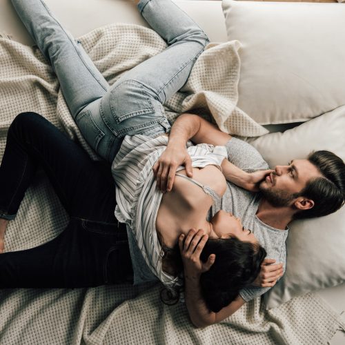 A couple lies on a bed, cuddling closely. The man is on his back, gently holding the woman, who rests her head on his chest. They appear relaxed and comfortable, surrounded by pillows and a light blanket.