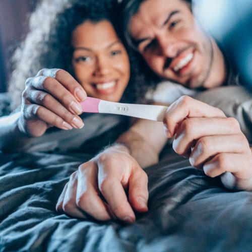 A smiling couple lies on a bed, holding a positive pregnancy test together, expressing excitement and happiness.