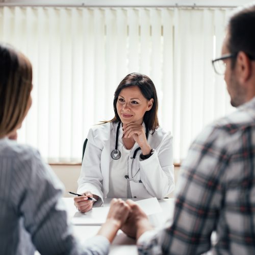 A doctor in a white coat sits at a desk, smiling and talking with a couple who are holding hands. The doctor appears to be giving advice or consultation in a bright office with vertical blinds.