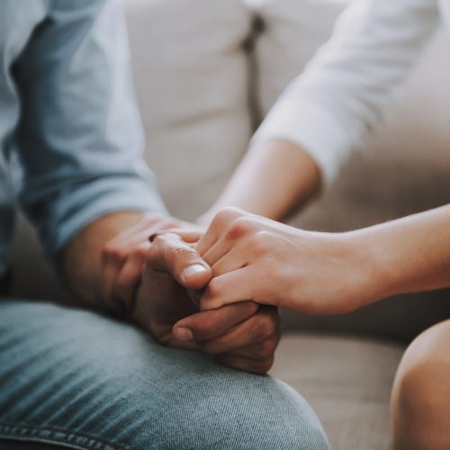 Two people sitting close together on a couch holding hands, one person gently grasping the others hand in a supportive or comforting gesture. Both are wearing casual long-sleeved shirts.