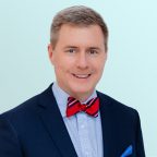 A man in a navy suit with a light blue shirt and a red striped bow tie smiles at the camera against a light blue background.