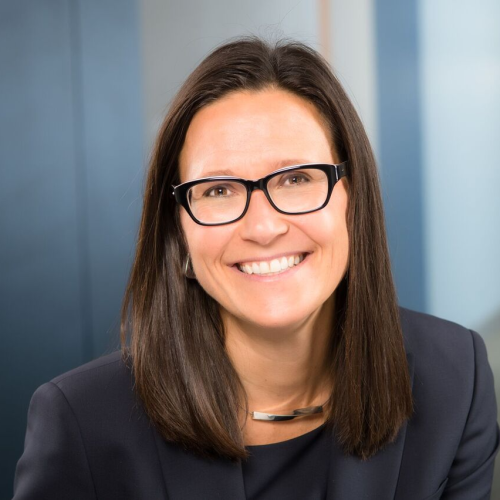A woman with straight brown hair and black glasses smiles at the camera, wearing a dark blazer and a silver necklace, with a blurred office background.