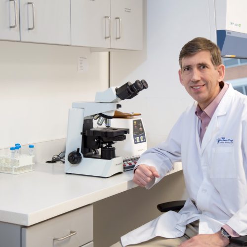 A man in a white lab coat sits at a laboratory desk next to a microscope, test tubes, and medical supplies, smiling at the camera inside a bright, modern laboratory.