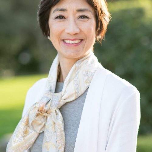 A woman with short brown hair smiles outdoors. She is wearing a white jacket, a grey top, and a patterned scarf tied around her neck. The background is blurred greenery and sunlight.