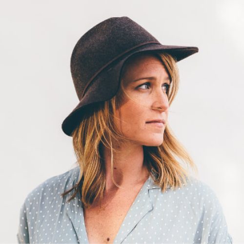 A woman with shoulder-length blonde hair wears a wide-brimmed brown hat and a light blue blouse with white polka dots, looking to her left against a plain light background.