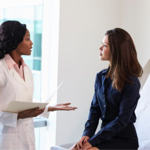 A doctor in a white coat speaks to a woman sitting on an exam table in a medical office, holding a file while the patient listens attentively.