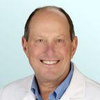 A smiling middle-aged man wearing a white lab coat over a light blue button-up shirt, standing against a light blue background.