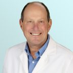 A smiling middle-aged man wearing a white lab coat over a light blue button-up shirt, standing against a light blue background.