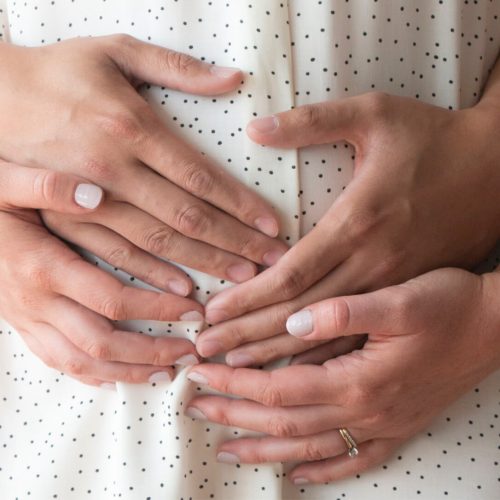 Close-up of four hands gently resting on a person’s belly, who is wearing a white shirt with black dots. Two hands are placed over two others, symbolizing care, support, or pregnancy.