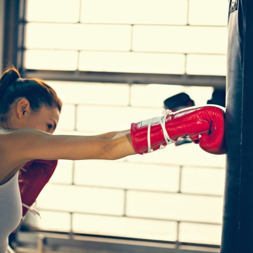 A woman wearing red boxing gloves and a white tank top punches a black punching bag in a gym, with sunlight streaming through large windows behind her.