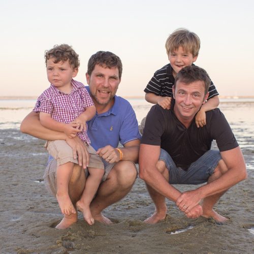 Two men and two young boys pose and smile together on a sandy beach during sunset. The men crouch down, with one boy sitting on each of their knees. The background shows calm water and a clear sky.