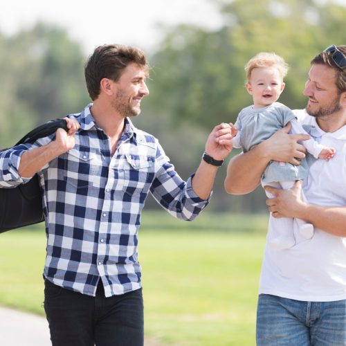 Two men stand outside on grass, smiling at each other. One is holding a baby, and the other carries a bag over his shoulder. The baby is also smiling. Trees and a blurred park background are visible.