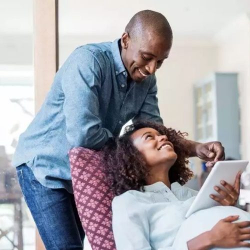 A smiling man leans over a seated pregnant woman who is holding a tablet. They are indoors, looking at each other affectionately, with the woman appearing relaxed and comfortable. Flowers and household items are visible in the background.
